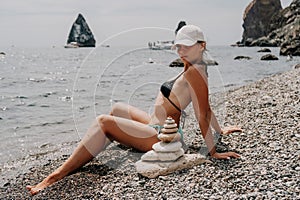 Woman bilds stones pyramid on seashore on a sunny day on the blue sea background. Happy holidays. Pebble beach, calm sea