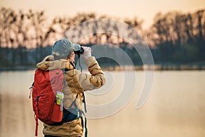 Woman with binoculars is looking for animals and birds at lake during sunset