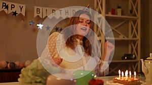 A woman with a birthday-cap on her head dances while sitting at a festive table. Candles are burning on the