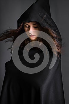 woman in a black cloak with a hood in the studio on a gray background. Long hair fluttering on the wind.