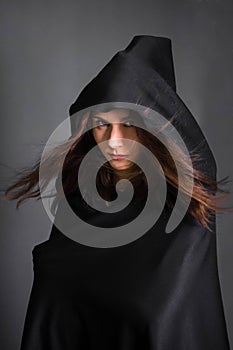 woman in a black cloak with a hood in the studio on a gray background. Long hair fluttering on the wind.