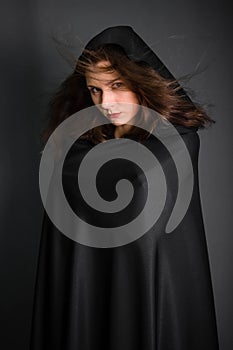 woman in a black cloak with a hood in the studio on a gray background. Long hair fluttering on the wind.