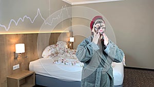 Woman with a black cosmetic mask in a towel on her head stands near the bed in a bathrobe, concept of facial treatment