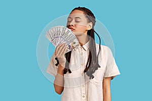 Woman with black dreadlocks holding big fan of money, smelling dollar banknotes with pleasure.