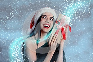 Woman in black dress and Santa hat holding Christmas gift on color background