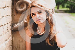 woman in black dress and white pearl necklace touching long hair, looking at camera and sitting near breek wall