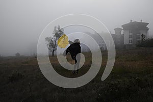 The woman in a black jacket walks with a yellow umbrella through the fog in forest.