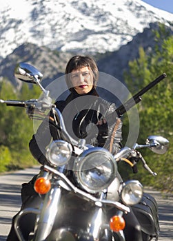 A woman in a black leather biker jacket with a carbine rifle on a chopper motorcycle in Greece on a road in the forest in the moun