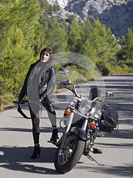 A woman in a black leather biker jacket with a carbine rifle on a chopper motorcycle in Greece on a road in the forest in the moun