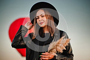 A woman in a black leather jacket and hat stands in front of a red balloon. Hot air balloon in flight in the sky