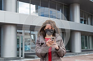 Woman with a black medical mask on her face with an anxious expression looks at smartphone screen against background of