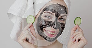 Woman with black purifying charcoal facial mask, holding cucumber slices covering her eyes