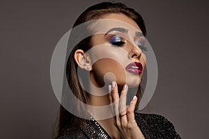 Woman in black shiny dress is touching her face, posing on gray background. Luxury makeup. Colorful eyeshadow, false