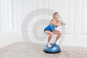 Woman in black shorts and blue top working in gym doing exersice in bosu balance trainer, squats on fitness ball, holding balance