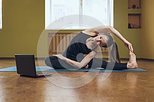 Woman in black sportswear does yoga, performs online exercise Parivritta Janu Shirshasana, looks into a laptop