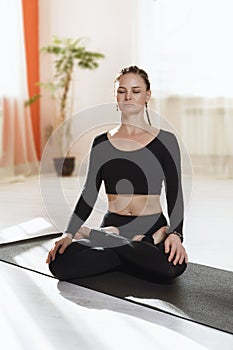 A woman in black sportswear practicing yoga sitting on a gymnastic mat in a lotus position is engaged in meditation at home