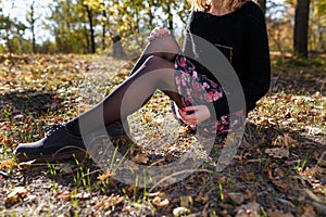 Woman in black sweater and sirt with flowrs sitting in autumn forest. Female hands with red manicure hugging legs