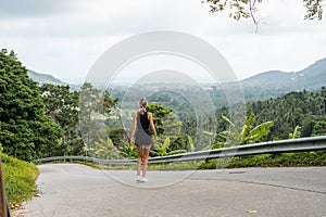 Woman in black t-shirt enjoying tropical forest view while standing on a empty road. Mountains and white clouds on a