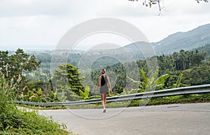 Woman in black t-shirt enjoying tropical forest view while standing on a empty road. Mountains and white clouds on a
