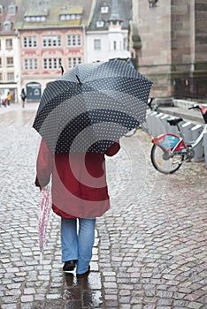 Woman with black umbrella and red coat walking in cobbles street