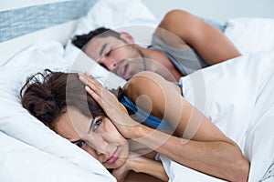 Woman blocking ears with hands while man snoring on bed