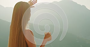 A woman blocks the sun with her hand, holding a tea cup. Golden hour light envelops female against mountainous outlines.