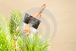 Woman blogger with laptop applying sunscreen on skin sitting under palm tree branches on the sand of beach . Summer vacation.