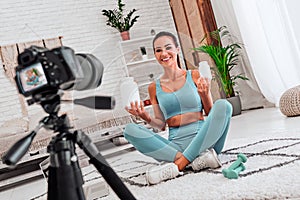 Woman blogger in sportswear sitting on the floor with dumbbells and a laptop and showing a jar of sports nutrition proteins to the