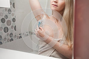 woman with blond hair and in a white towel shaves her armpits in the bathroom