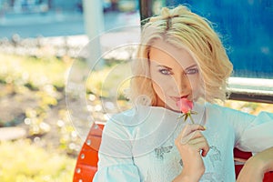 Woman blonde hair holding smelling a pink rose looking at you camera boyfriend husband sitting on red bench isolated city outdoors