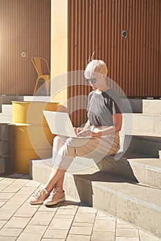 Woman with blonde hair working on laptop sitting outside at stairs, hide from the sun, sunny day, chill.