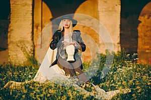 Woman blonde in hat sitting on chair in nature on background of old building with arches. Lady is holding an aquarium