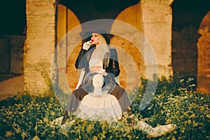 Woman blonde in hat sitting on chair in nature on background of old building with arches. Lady is holding an aquarium