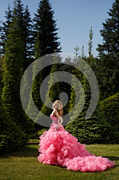 Woman with blonde locks wearing pink evening dress with fluffy skirt is posing in botanical garden on the grass