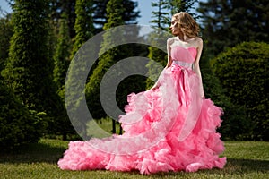Woman with blonde locks wearing pink evening dress with fluffy skirt is posing in botanical garden on the grass