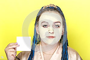 Woman with blue afro braids face in a frozen mask of green clay with a business card in her hands on a yellow background