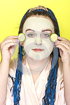 Woman with blue afro braids face in a frozen mask of green clay with pieces of cucumbers in her hands on a yellow background