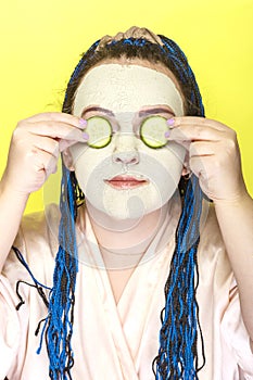 Woman with blue afro braids face in a frozen mask of green clay with slices of cucumbers in front of her eyes on a yellow