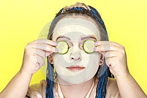 Woman with blue afro braids face in a frozen mask of green clay with slices of cucumbers in front of her eyes on a yellow