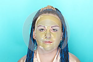 Woman with blue afro braids face in a mask of green clay on a blue background.