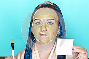 Woman with blue afro braids face in a mask of green clay on a blue background with a brush in her hands