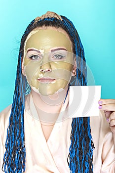 Woman with blue afro braids face in a mask of green clay on a blue background with a brush in her hands and a business card