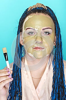 Woman with blue afro braids face in a mask of green clay on a blue background holds a brush in her hands