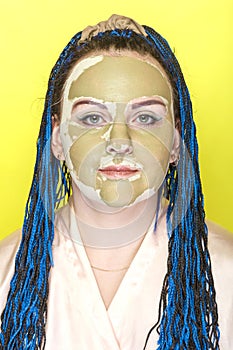Woman with blue afro braids face in a mask of green clay on a yellow background