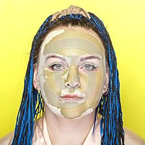 Woman with blue afro braids face in a mask of green clay on a yellow background