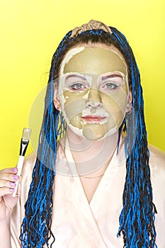 Woman with blue afro braids face in a mask of green clay on a yellow background with a brush in her hands.