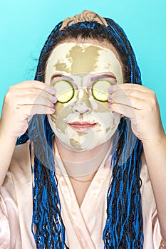 Woman with blue afro braids face in a mask made of green clay with cucumber circles on her eyes on a yellow background.