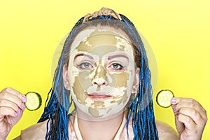 Woman with blue afro braids face in a mask made of green clay with cucumber circles in her hands on a yellow background