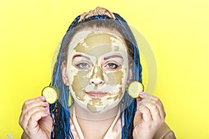 Woman with blue afro braids face in a mask made of green clay with cucumber circles in her hands on a yellow background