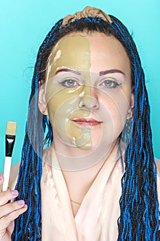 A woman with blue afro braids, half of her face in a mask of green clay on a blue background holds a brush.